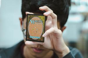 Male with black hair and glasses holding Magic the Gathering Card infront of his face. 